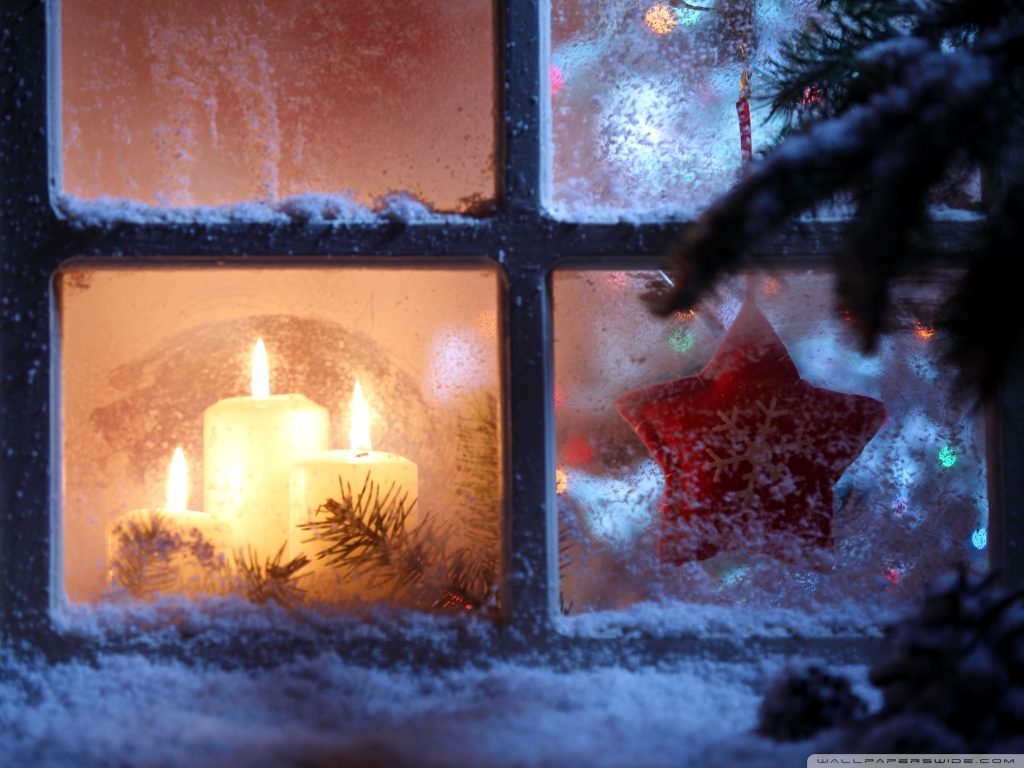 A view of a snowy window, behind which are three burning white candles and a red star-shaped ornament.