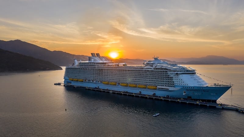 An ocean liner sails out to sea at sunset, with a mountain in the background.