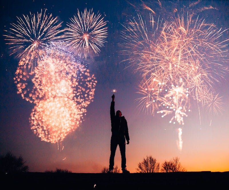 A man stands with a sparkler in silhouette in front of a beautiful New Year's Eve fireworks display.