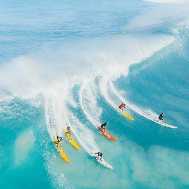 Clear blue sea in Hawaii with rising wave, six surfers ride it.